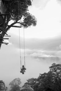 Rear view of woman sitting on swing hanging from swing against cloudy sky