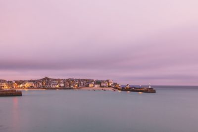 Illuminated city by sea against sky at sunset