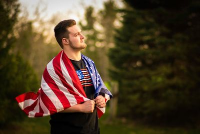 Thoughtful man holding american flag while standing against trees