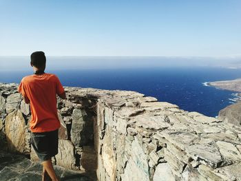 Rear view of man standing on cliff by sea against clear sky