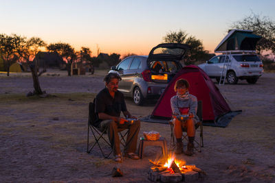 Couple sitting bonfire on field against sky during sunset