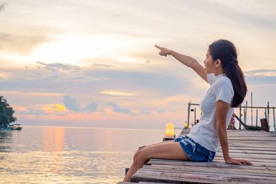 Woman sitting on pier over sea against sky during sunset