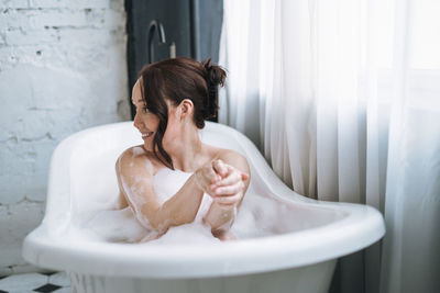 Self loving adult smiling woman with brunette hair taking bath with foam at home