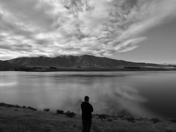 Rear view of silhouette man standing by lake against cloudy sky