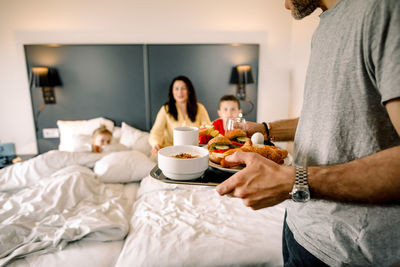 Father holding breakfast with family sitting on bed in background