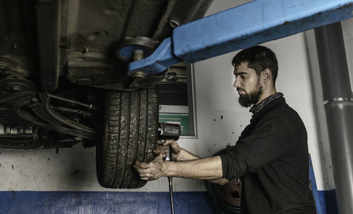 Side view of male master using electric screwdriver while fixing wheel of automobile