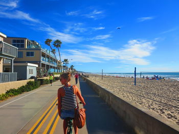 Rear view of woman riding bicycle on walkway by beach against blue sky