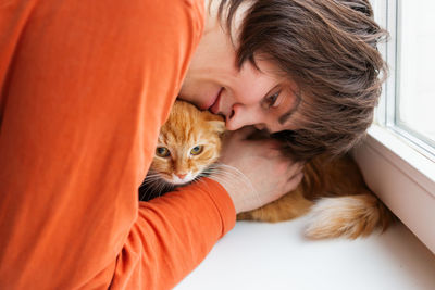 Man in orange sweatshirt is hugging cute ginger cat. pet adoption. ex-stray cat at home. fluffy pet.