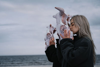 Woman holding cow skull by the sea against cloudy sky