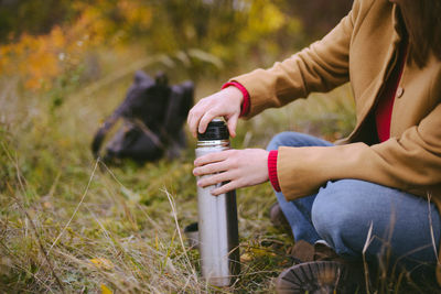 Low section of woman holding insulated drink container while sitting on field during autumn