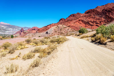 Dirt road by rocky mountains against sky on sunny day