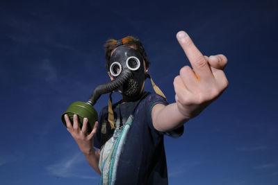 Boy wearing gas mask while showing obscene gesture against sky