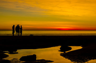 Silhouette male friends standing at beach against orange sky