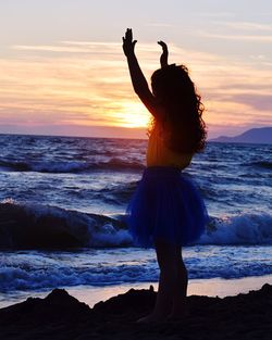 Rear view of girl with arms raised standing at beach against cloudy sky during sunset