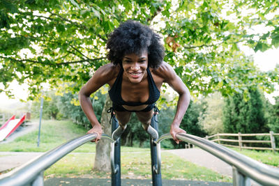 Low angle of positive young curly haired black sportswoman doing push ups on parallel bars in summer park