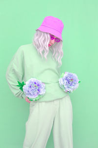 Woman wearing hat holding flowers standing against colored background
