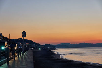 Illuminated street by sea against sky during sunset