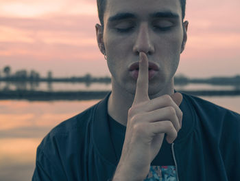 Close-up of young man with finger on lips by lake during sunset