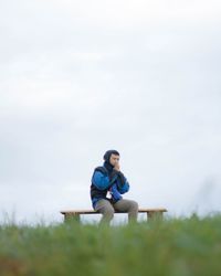 Young man sitting on bench against sky