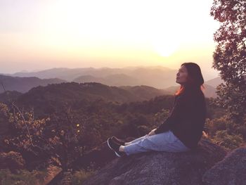 Woman sitting on rock against sky during sunset