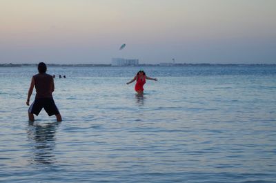 Friends playing with plastic disc while standing in sea against sky during sunset