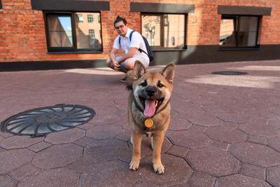 Mid adult man looking at dog panting while standing on road against building