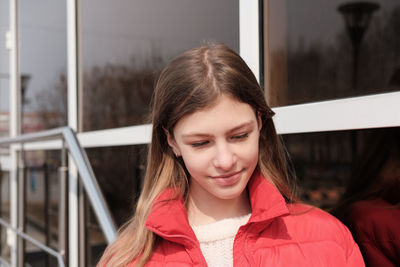 Happy smiling teenager girl in winter jacket standing against window wall. street portrait 