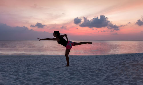 Full length of woman practicing yoga at beach against cloudy sky during sunset