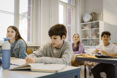 Schoolboy writing in book while sitting at desk with friends in classroom