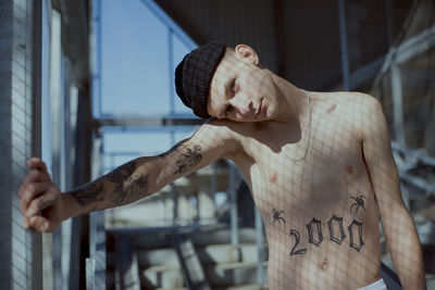 Portrait of shirtless young man with tattoo standing indoors