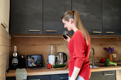 A young girl with a glass of wine in the kitchen is looking at a digital tablet for a salad recipe 