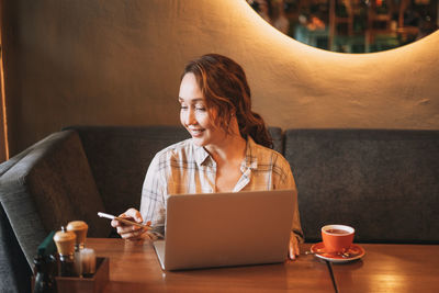 Adult charming brunette woman in plaid shirt working with laptop using mobile phone at cafe