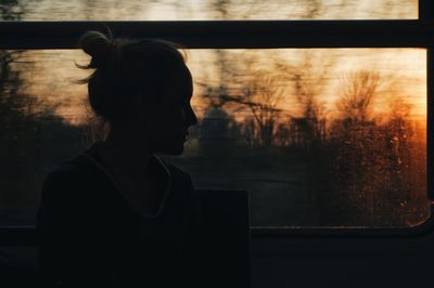 Woman sitting against window in train during sunset