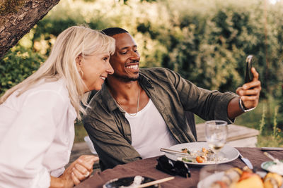 Happy man taking selfie with mother while sitting at dining table in back yard