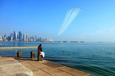 Man looking at air show while sitting on promenade by city against clear blue sky