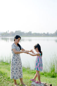 Full length of mother and daughter standing at riverbank against clear sky