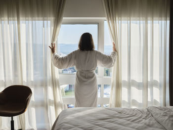 Woman opening white curtain while looking through window at luxury hotel room