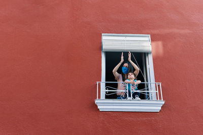 Masked mother clapping from the window with her little daughter on a red background