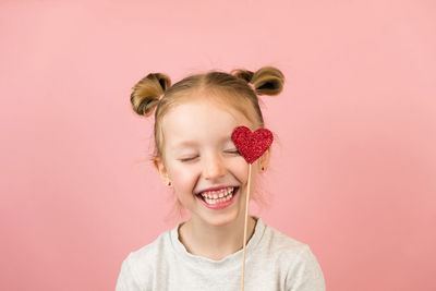 Funny little blonde girl smiling and playing with red heart toy on pink background