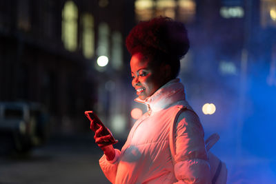 Smiling black girl hold smartphone look at screen reading message waiting for friends at night city