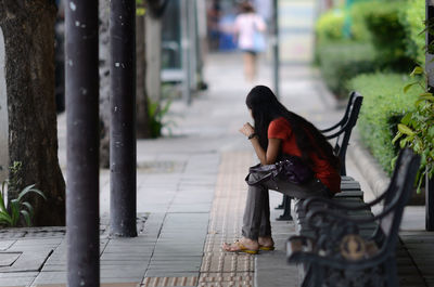 Full length of woman sitting on bench at sidewalk