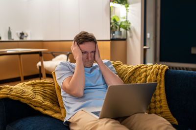 Tired of work on laptop man touch head, tries to concentrate looks at screen sitting on sofa at home