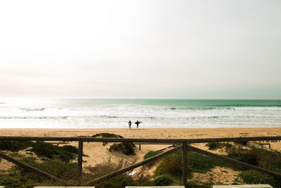 Distant view of friends with surfboard standing at beach against sky during sunny day