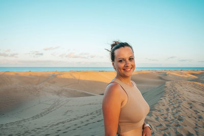 Portrait of young woman standing at beach against sky during sunset