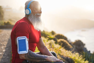 Side view of smiling bearded man listening music while sitting on mountain