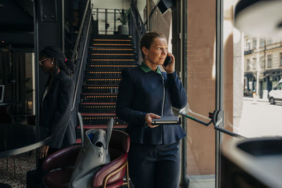 Mature businesswoman holding diary while talking on smart phone by female colleague in hotel lounge