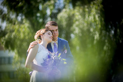 Portrait of bridegroom with bride standing against trees