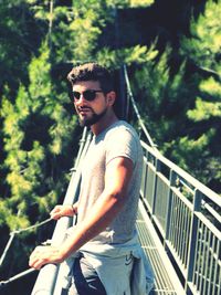 Young man wearing sunglasses standing by railing against trees