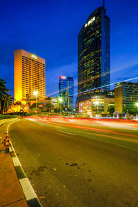 Light trails on city street by buildings against sky at night