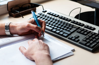 Cropped hands of businessman writing in book by keyboard on office desk